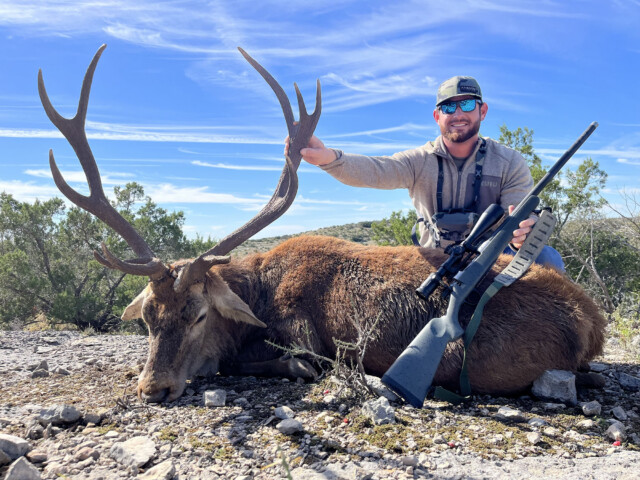 Hunter kneeling beside a large elk with massive antlers on rocky desert ground, rifle resting nearby under a blue sky.