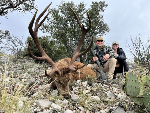 Two hunters pose beside a large elk with massive antlers on a rocky desert terrain, smiling at the camera.