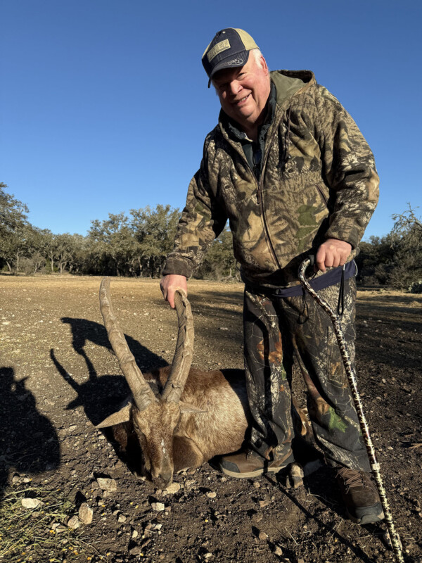 Smiling hunter in camo outdoors kneels beside a large ram with curling horns on a rocky dirt ground under a blue sky.